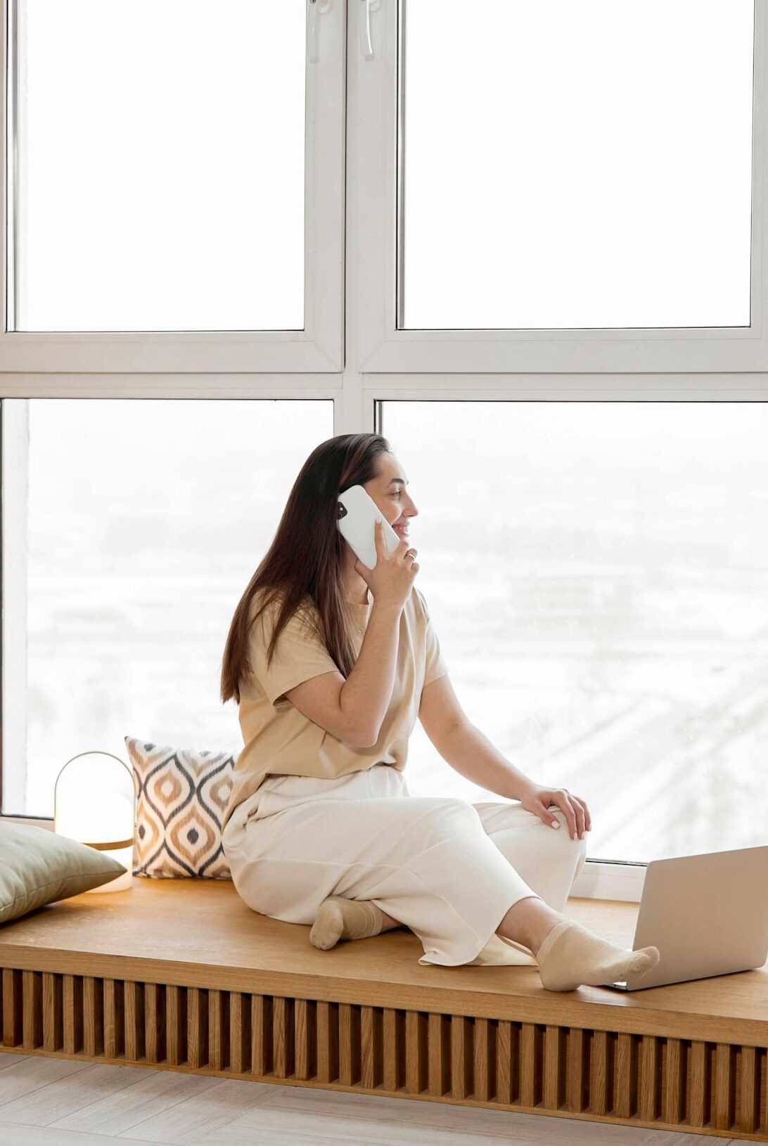 A woman talking on a smartphone while sitting by a large window with a laptop. A cozy indoor setting.