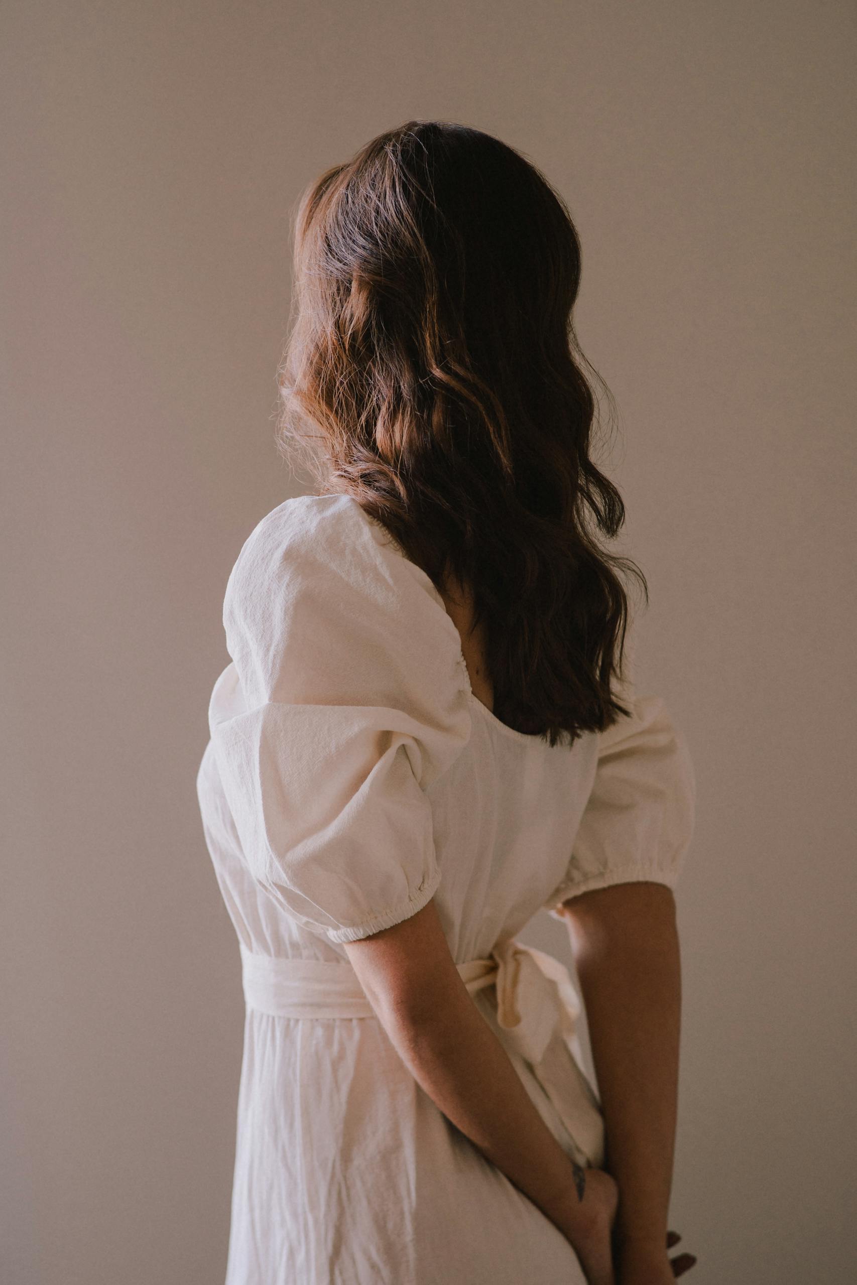 Back view of a woman in a chic white dress posing indoors with gentle light.